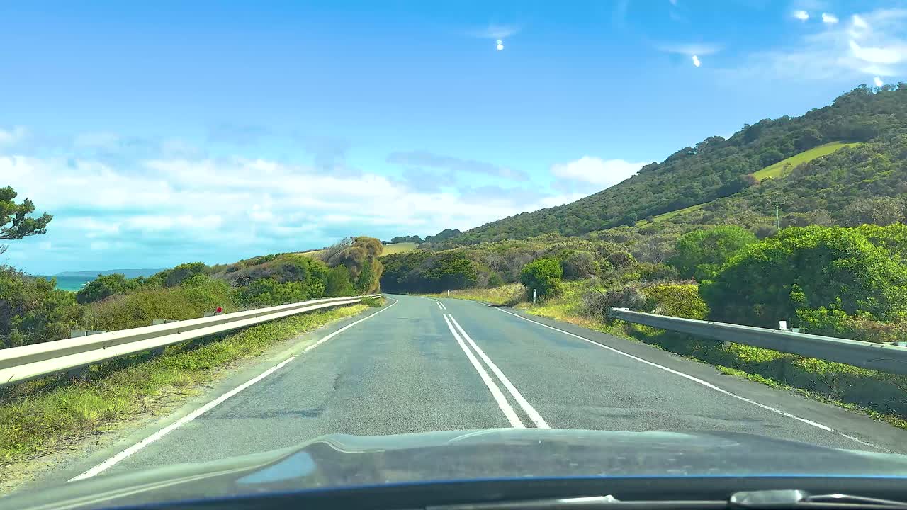 A car journey on a sunny day along the winding Great Ocean Road, surrounded by lush greenery and coastal views