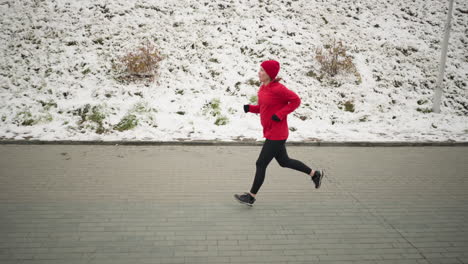 woman jogging outdoors on interlocked pavement next to snowy hill during winter, dressed in vibrant red hoodie and black leggings, scene showcases serene natural hillside with snow