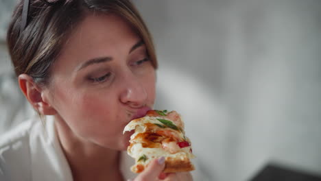 close up of calm woman eating delicious pizza slice while glancing thoughtfully at something distant in softly blurred background, wearing white clothing with eyeglasses resting on head