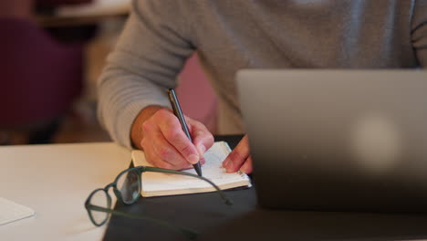 mature businessman working on laptop at desk in office making notes in notebook