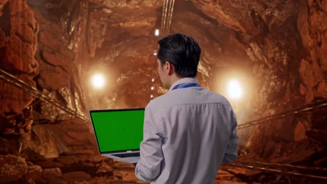 back view of an asian male professional worker standing in underground mine tunnel, typing on laptop's keyboard with green screen and look at the factory once