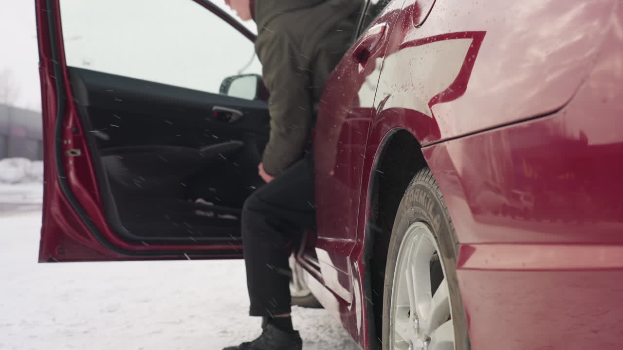 man in winter jacket exits red car during heavy snowfall in snowy parking area near industrial building, wet glossy surface glistens with droplets, background shows parked vehicles