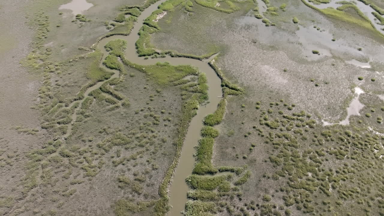 Aerial video of a winding tidal creek cutting through the marshland of Hilton Head Island. The waterway meanders through dense patches of grass and muddy inlets, forming organic patterns
