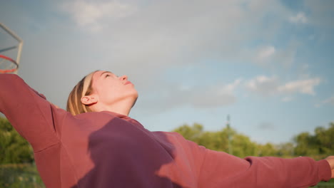 young lady in hoodie serving volleyball outdoors with basketball hoop visible in background, preparing for serve in open court with a calm, focused expression and natural daylight