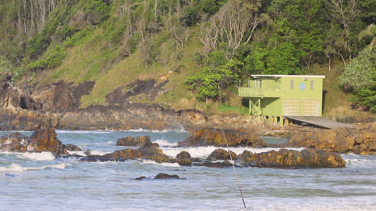 Ocean waves crash against rocky shore near a green building, under soft daylight at Charlesworth Bay Beach, Coffs Harbour