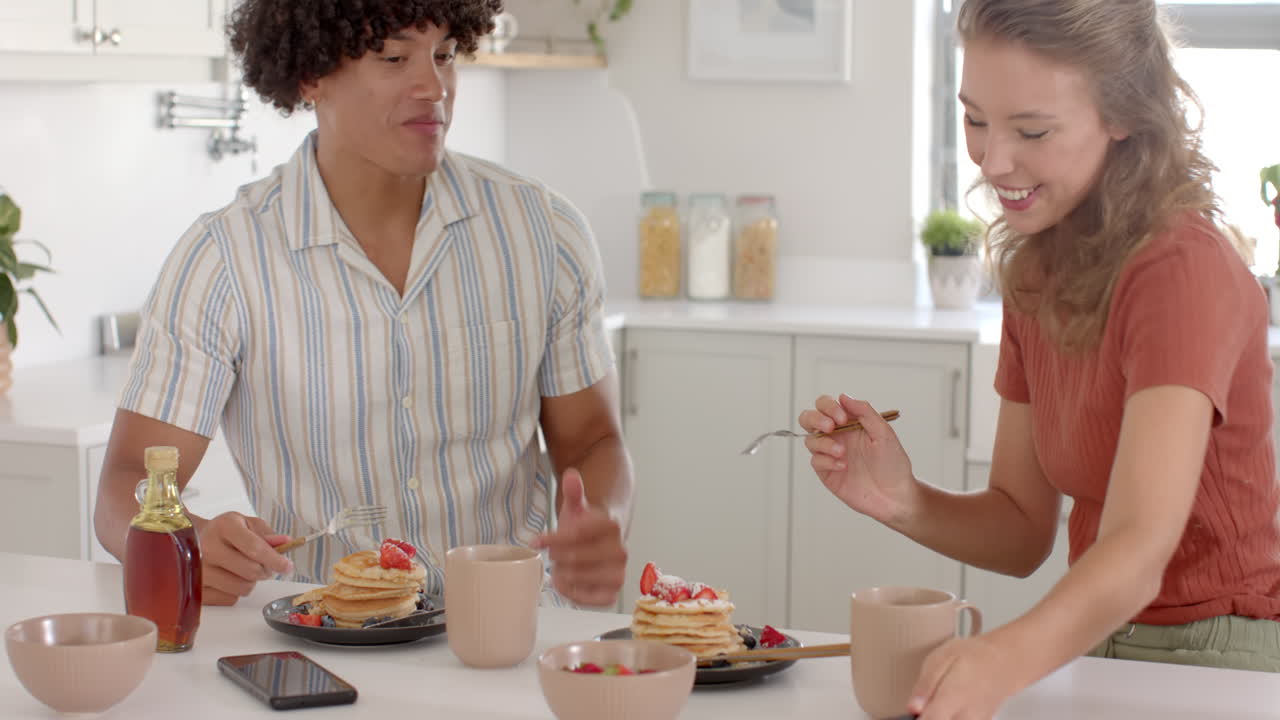 Sharing breakfast, diverse couple having pancakes and coffee, laughing together at home