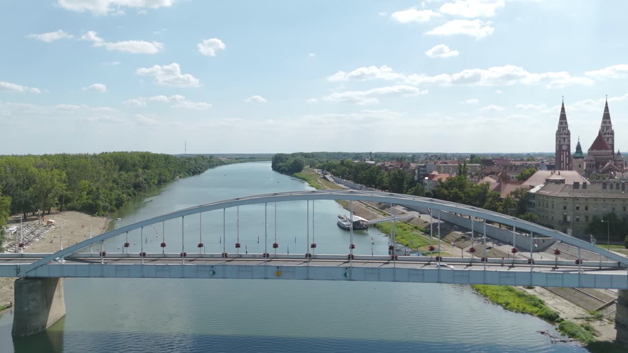 Smooth backward drone flight revealing Szeged’s Belvárosi Bridge spanning the Tisza River, with the majestic Votive Church rising in the background