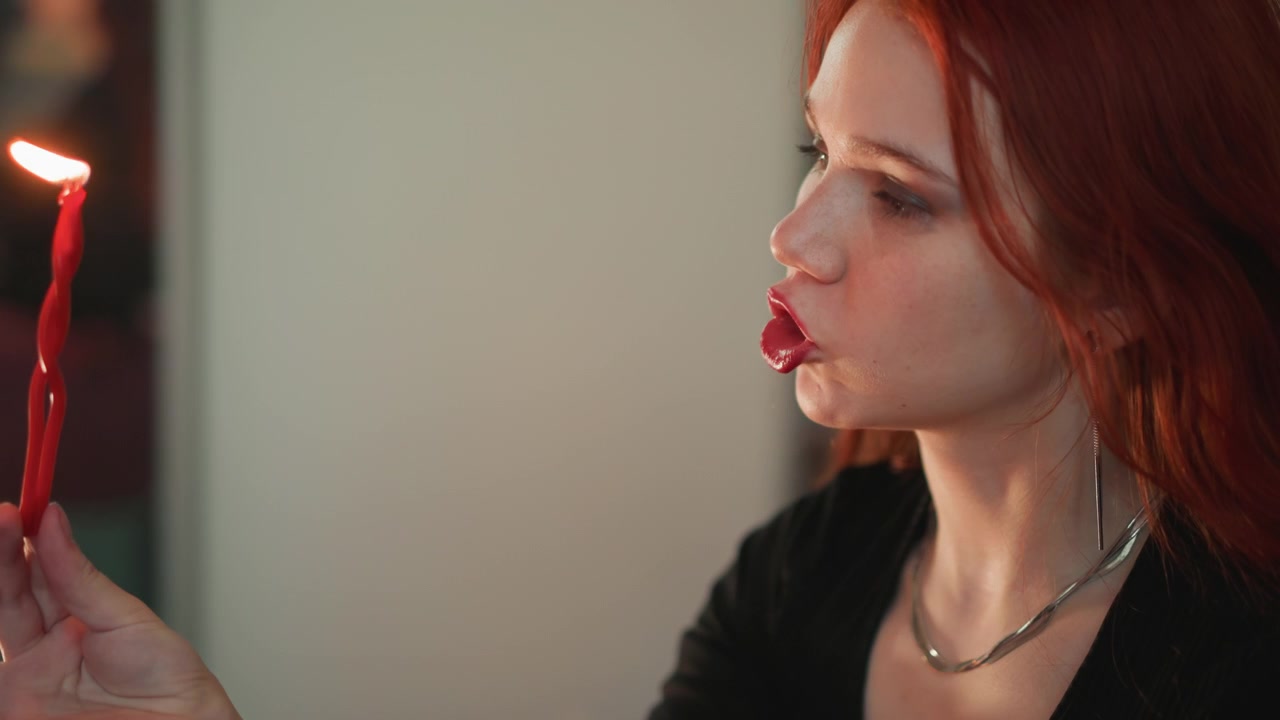 Observer watches carefully. Redhead watches dim room ritual. Woman with red hair observes candle ritual attentively. Brunette closely watches flickering ceremony within dimly lit space