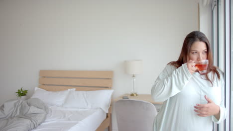 plus size biracial woman drinking tea looking out of bedroom window, copy space, slow motion