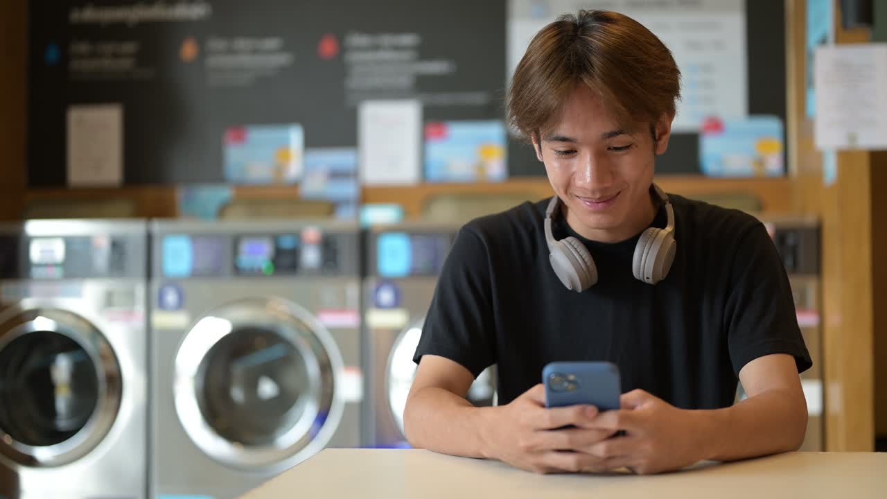 Young handsome Thai man sitting in a modern launderette with washing machines using phone, wearing a black t-shirt. Candid portrait showing Gen Z lifestyle, daily chores, laundry, and urban living