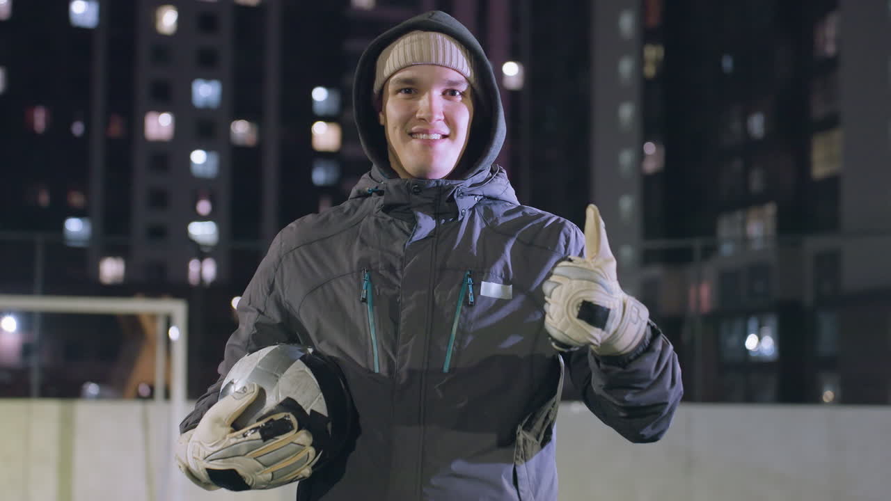 atleta sonriente con capucha y guantes alterna la pelota de fútbol en las manos y da pulgares hacia arriba con la mano izquierda durante el entrenamiento nocturno en el campo deportivo urbano rodeado de edificios iluminados de la ciudad