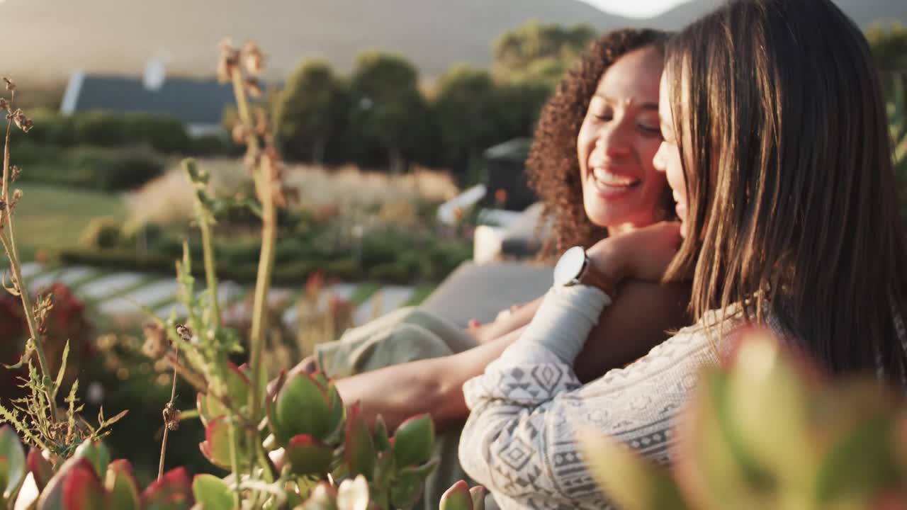 feliz pareja romántica de lesbianas biraciales sentada y abrazándose en el jardín al atardecer, cámara lenta