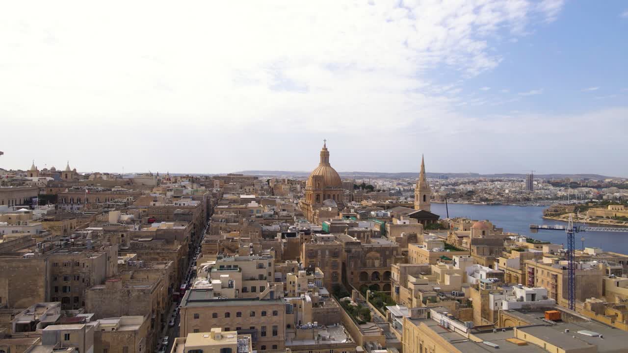 Ripresa aerea ravvicinata da drone che sorvola i fitti tetti in calcare dorato, le cupole di chiese barocche e le strette vie della storica La Valletta, Malta, con il porto blu visibile sullo sfondo