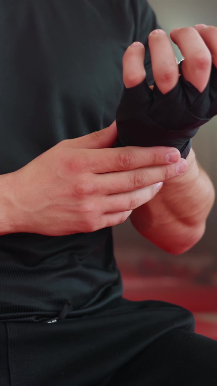 Close up of man in black clothing wrapping his hands. Background features red flooring consistent throughout. Soft lighting highlights skin tone contrast against dark attire