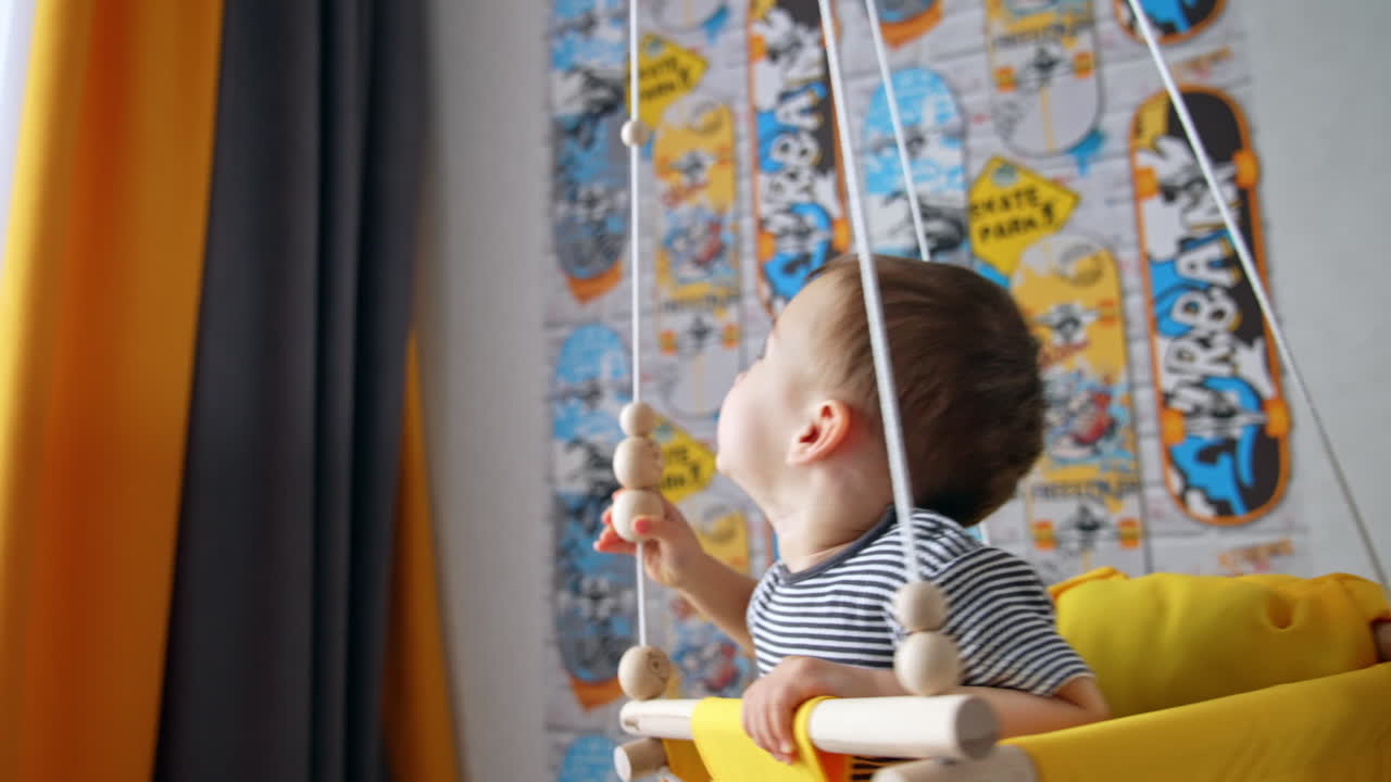 Little boy swings in a yellow swing indoors. Kid plays with wooden beads on the rope.