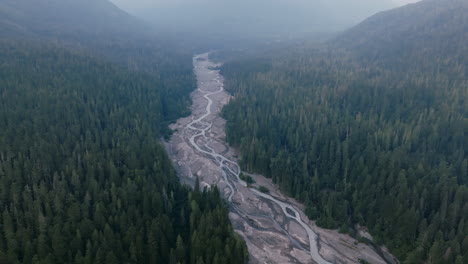 Slow-aerial-footage-in-the-hazy-morning-of-a-riverbed-that-is-in-between-a-forest-of-evergreen-trees-in-the-Cascade-Mountains