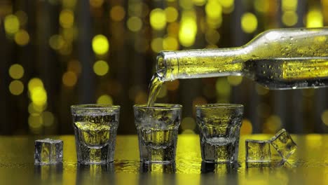 barman pour frozen vodka from bottle into shot glass. ice cubes against shiny gold party background