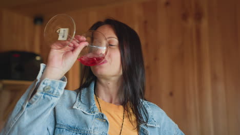 young brunette in denim jacket slowly sips red wine from crystal glass inside cozy wooden cabin kitchen, enjoying relaxed afternoon, eyes half closed savoring aroma amid rustic interior lighting