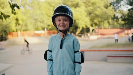 Portrait-of-a-happy-blond-boy-in-a-blue-jacket-in-a-black-helmet-and-elbow-pads,-who-stands-against-the-backdrop-of-a-skatepark-in-the-park