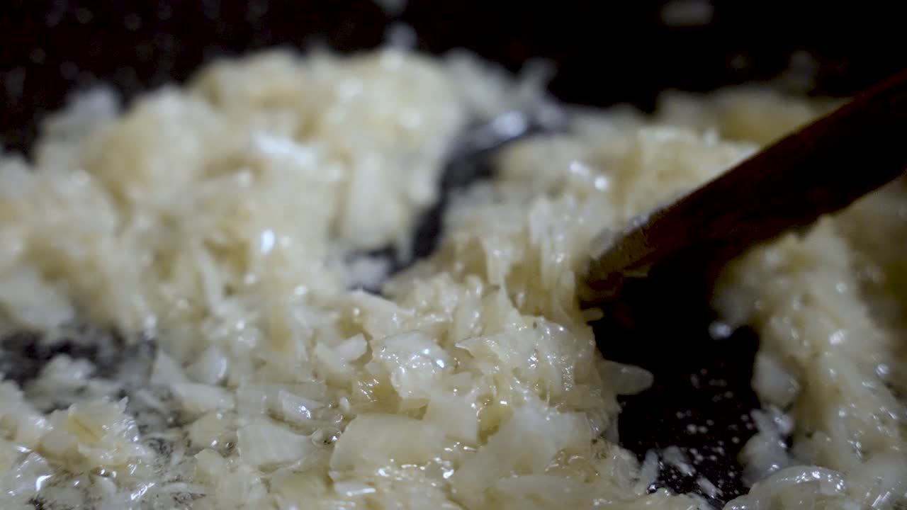 Chopped white onions are sizzling in hot oil in a pan while being stirred by a wooden spoon. slow motion
