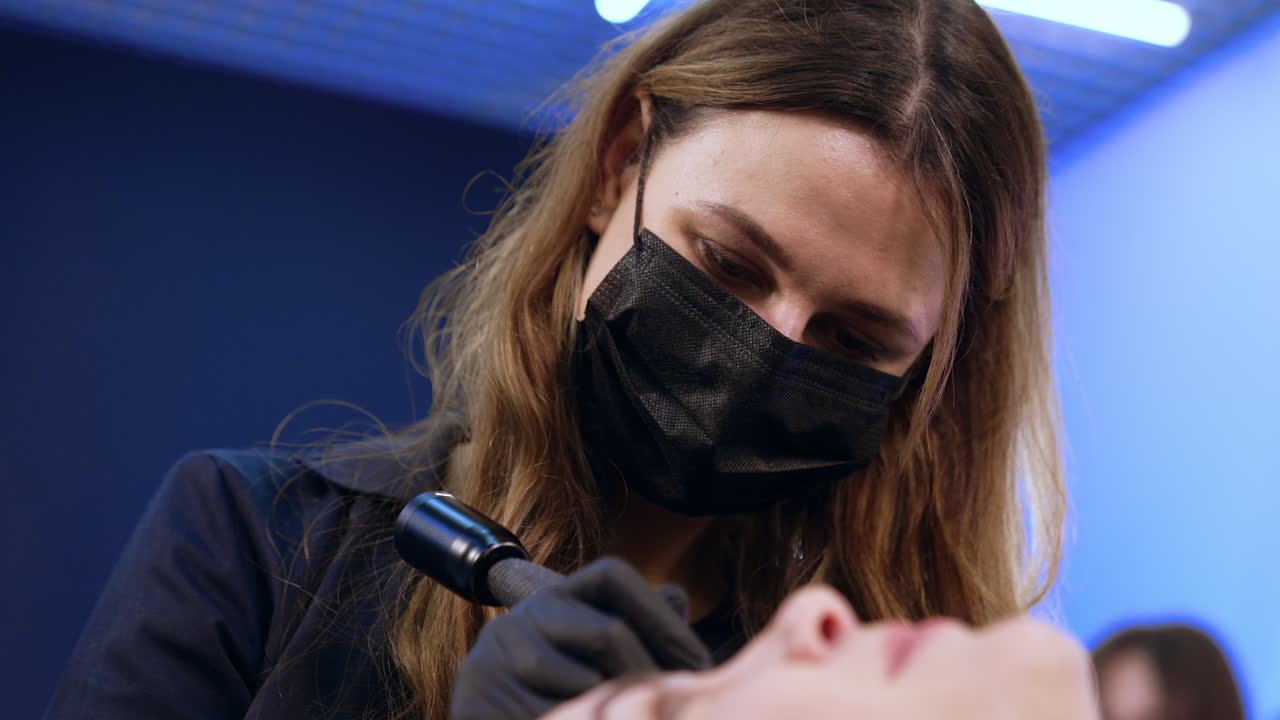 Long haired Caucasian beautician wearing black mask. Cosmetologist does permanent make up of eyebrows. Close up.