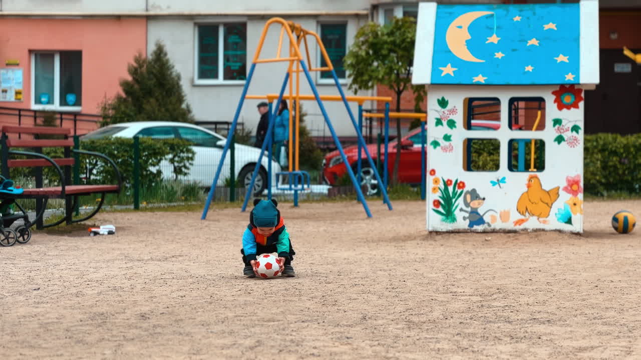Happy Caucasian toddler runs by the playground. Kid picks up a ball from and approaches the camera.
