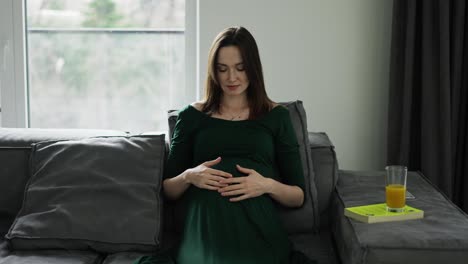 Portrait-of-beautiful-pregnant-woman-sitting-on-sofa-and-gently-touching-her-belly