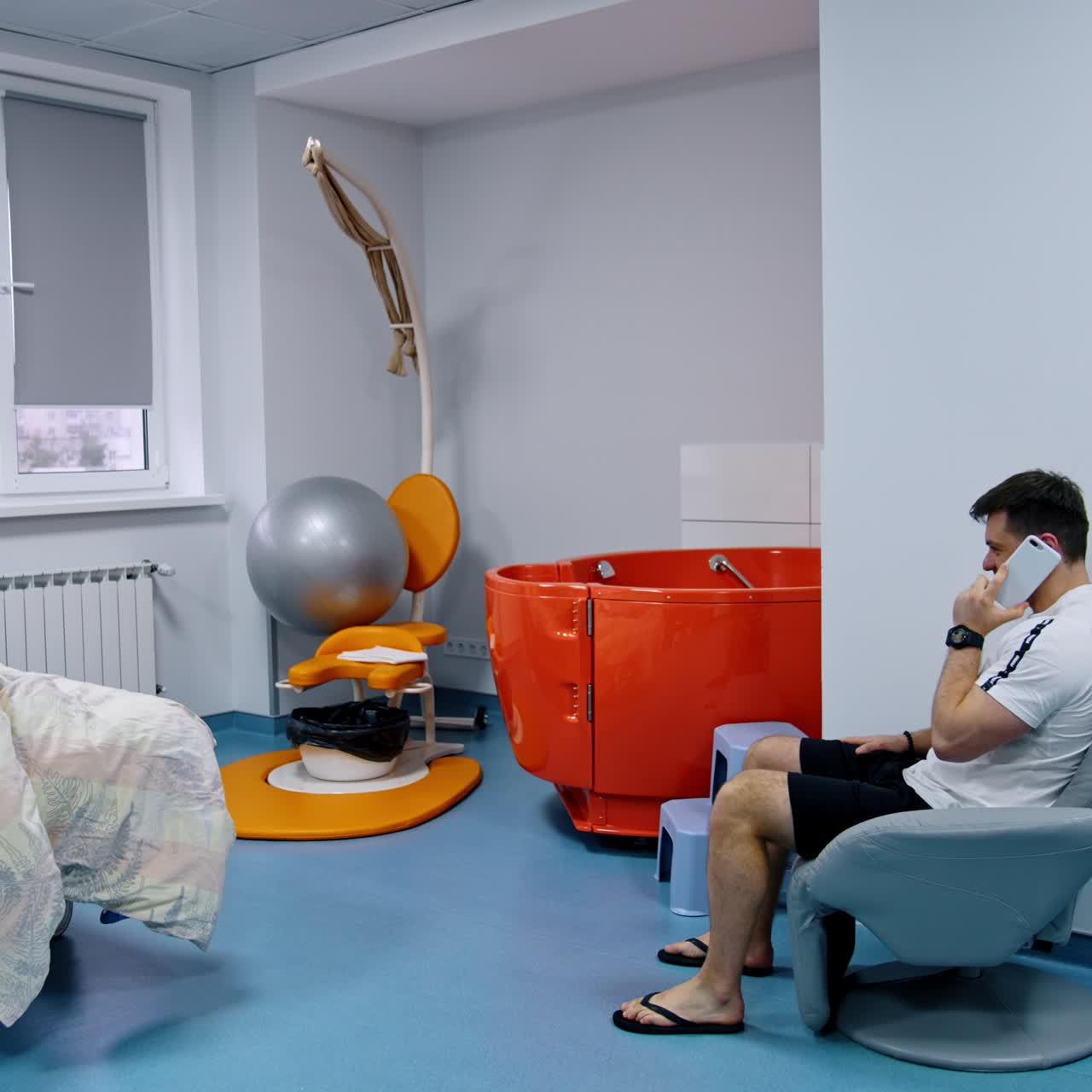 Pregnant Caucasian woman lies in the bed at maternity hospital. Husband sits in an armchair speaking on the phone
