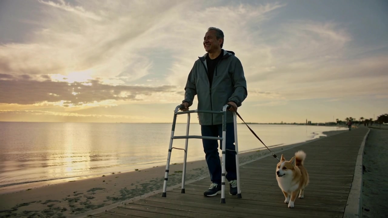 Corgi som drar i båndet, eldre mann som bruker rullator og går langs strandpromenaden i hettegenser ved solnedgang.