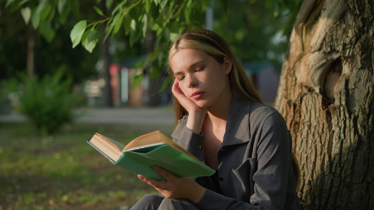Lady seated under tree with head resting on right hand, flipping through a book and appearing moody, sunlight gently touches her face while the soft, blurred background features distant objects