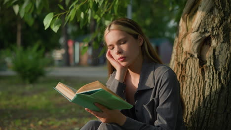 dama sentada bajo un árbol con la cabeza apoyada en la mano derecha, hojeando un libro y apareciendo de mal humor, la luz del sol toca suavemente su cara mientras el fondo suave y borroso presenta objetos lejanos