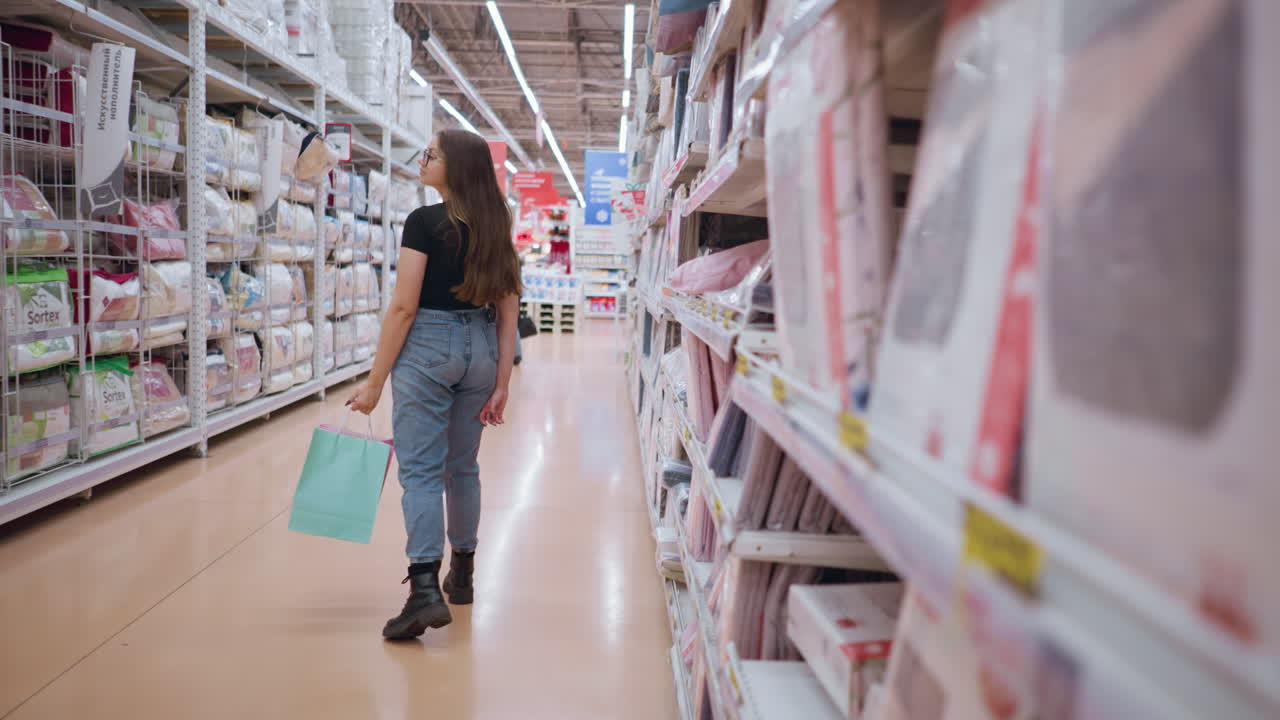 vista trasera de una mujer caminando por el pasillo del supermercado cerca de productos textiles, centrándose en las mercancías expuestas en los estantes mientras sostiene bolsas de compras, con otro comprador caminando delante