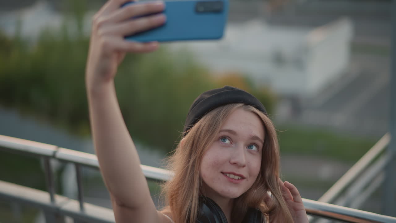 Selfie em close-up de uma mulher branca no terraço, usando fones de ouvido e gorro, capturando expressões espontâneas durante o pôr do sol em um mirante urbano, fundo da cidade com bokeh suave, luz quente natural, olhar divertido e sorriso sutil