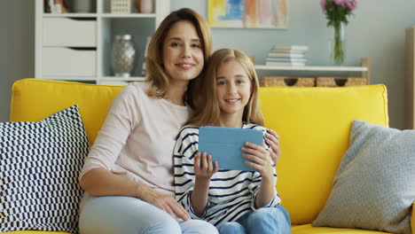 Madre-Rubia-Con-Su-Hija-Adolescente-Viendo-Videos-En-Tableta,-Hablando-Y-Sonriendo-Mientras-Están-Sentados-En-El-Sofá-En-La-Sala-De-Estar