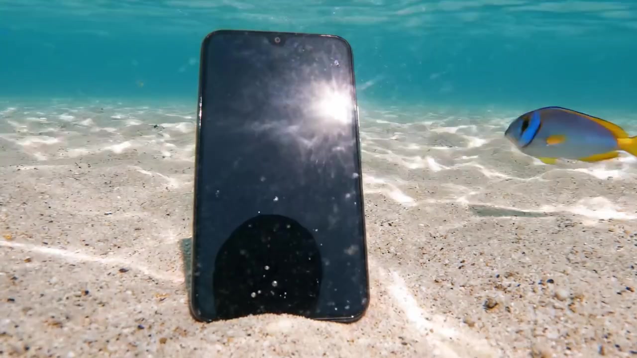 A smartphone rests on the sandy ocean floor, partially submerged in clear water, showcasing reflections and the serene underwater environment surrounding it.