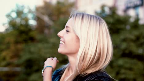 young blonde woman standing in the park and looking at camera. girl seriously looks and then starts to smile, laugh