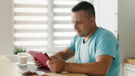 Junger-Mann-Liest-Nachrichten-Auf-Einem-Digitalen-Tablet-Am-Fenster-In-Seiner-Küche