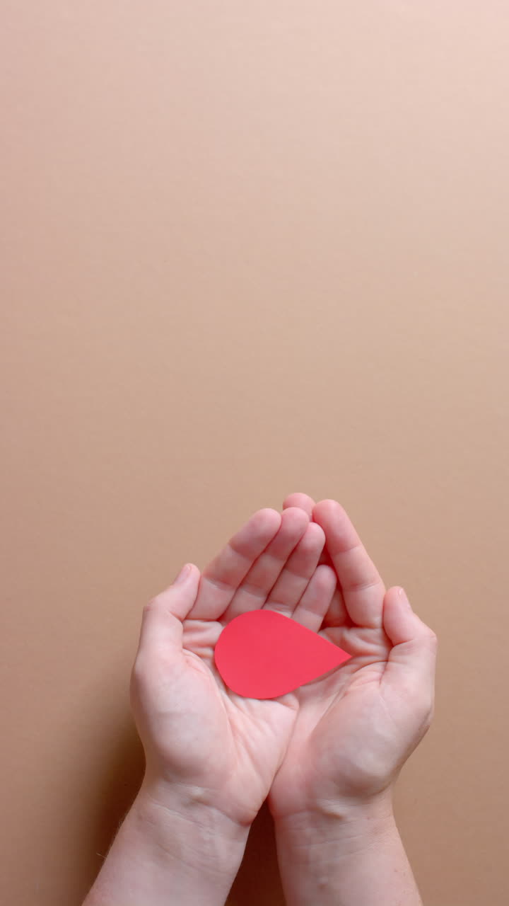 Vertical video of hands of caucasian man holding blood drop on beige background with copy space