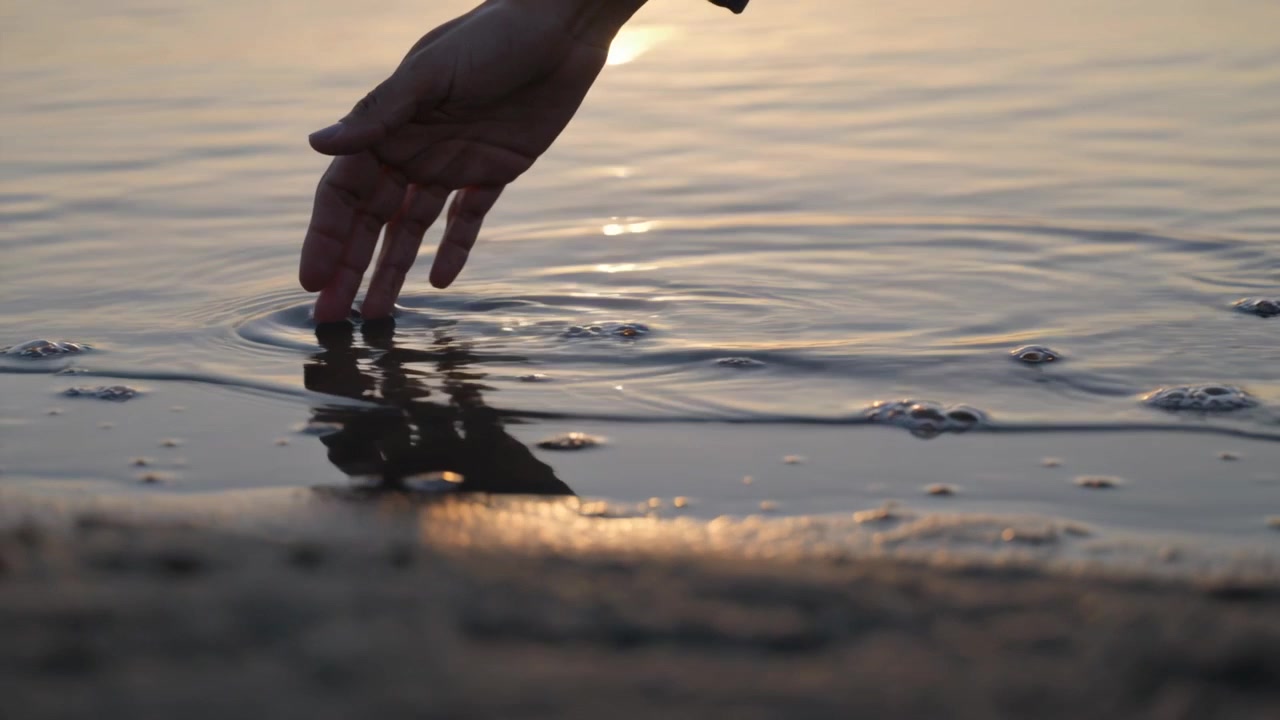 Sunlight glinting, female hand in dark sleeve reaching, touching wet sand and sea, creating ripples