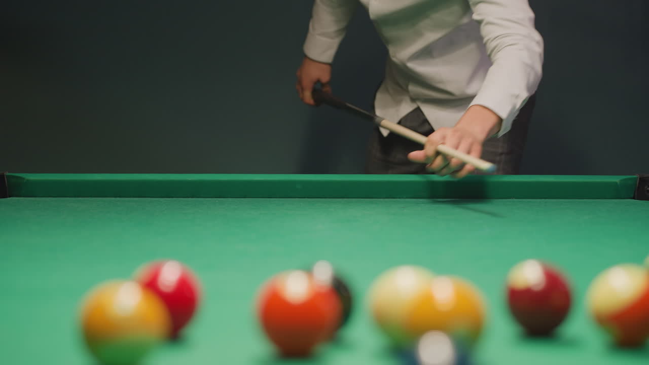 Young man in light shirt and dark trousers leans over green pool table, gripping cue stick with deep concentration. Eyes locked on balls, posture steady, creating tension in dimly lit game room