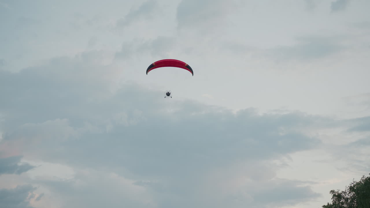 people flying powered paraglider under pastel sky with dramatic clouds and treetop silhouette, red canopy arching over horizon, motion captured in dusk light