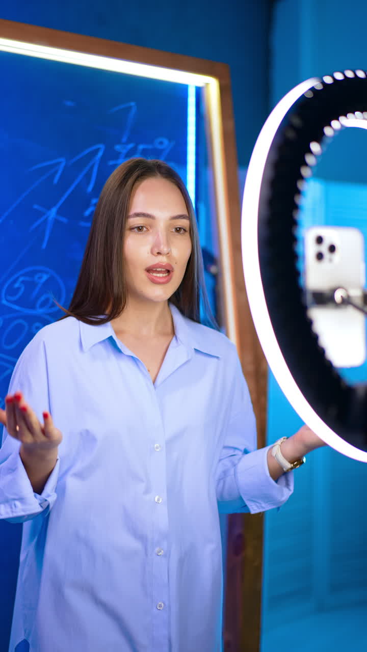 Caucasian girl wearing a blue shirt stands in front of phone on tripod and ring light. Confident lady talks and gestures with her hands. Vertical video.