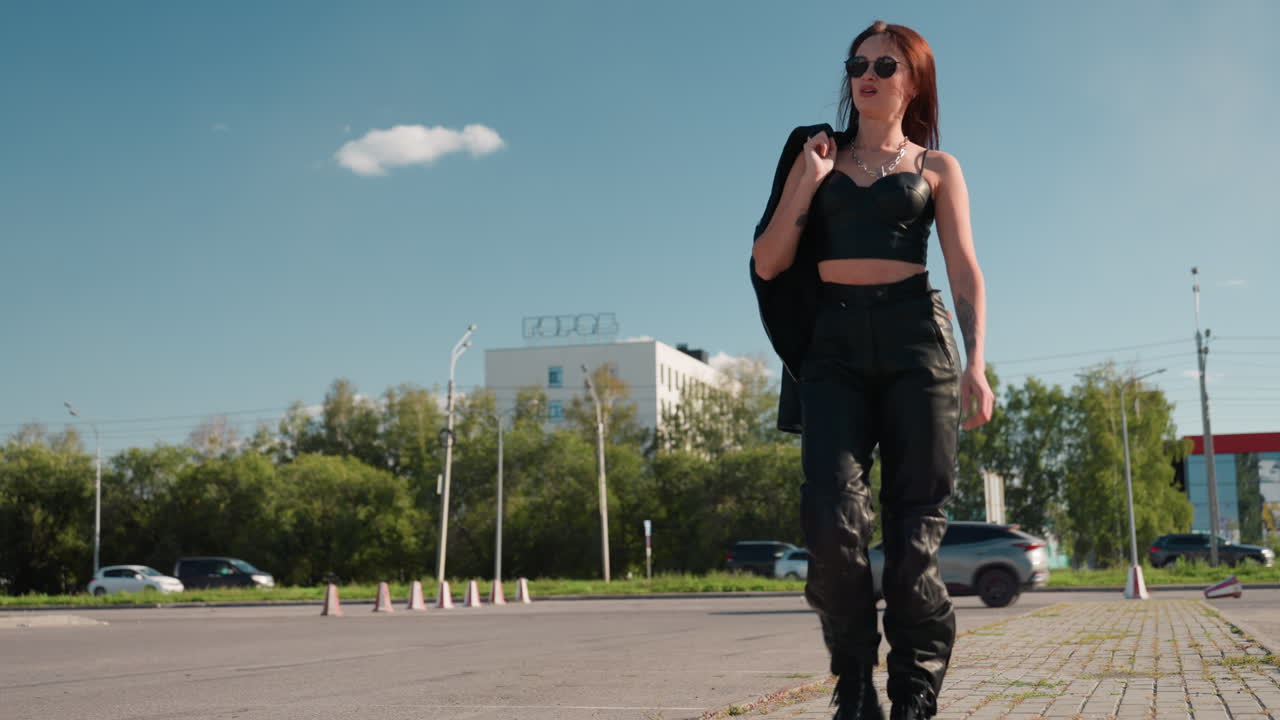 Lady walking confidently on interlocked pavement under blue sky wearing sunglass casually carrying jacket over shoulder with relaxed stride urban background of trees cars and buildings