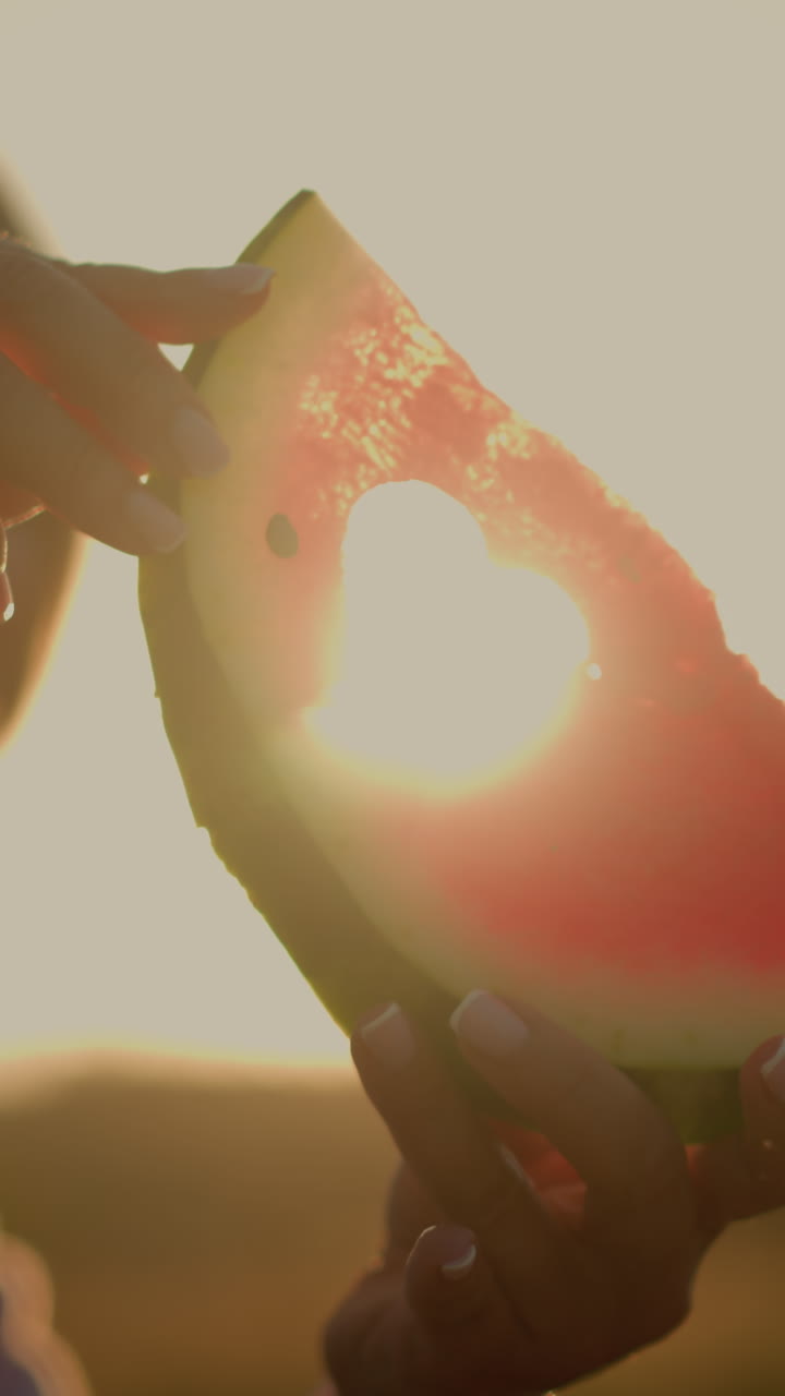 Close-up of woman holding watermelon slice with heart cutout shape against bright sunlight, illuminating vibrant red fruit and creating a warm, romantic silhouette