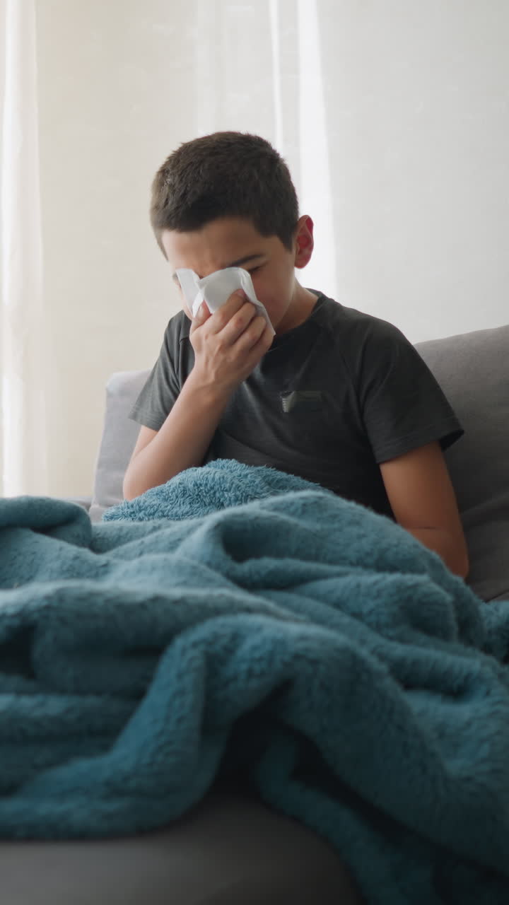 Young kid sneezing into tissue seated on couch under blue blanket, with tissue roll nearby and glass cup on the table, showing signs of sickness and fatigue, taking care of his health indoors