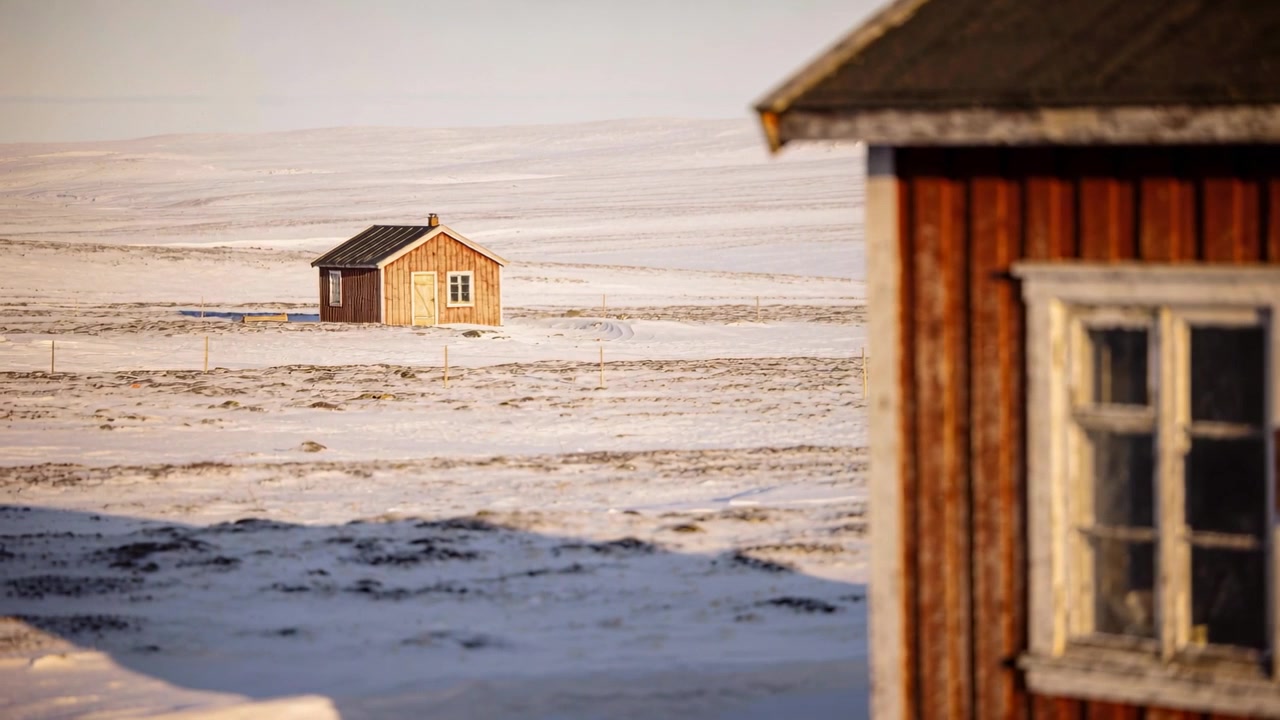 Panorerende kamera sentrerer en brun hytte på en snødekt slette, viser en rød bygning til høyre, med plass til tekst.