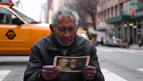 an elderly man sits on a busy city street, gazing thoughtfully at a photo of two men. the surrounding urban environment is bustling with traffic and life, contrasting his quiet moment of reflection.