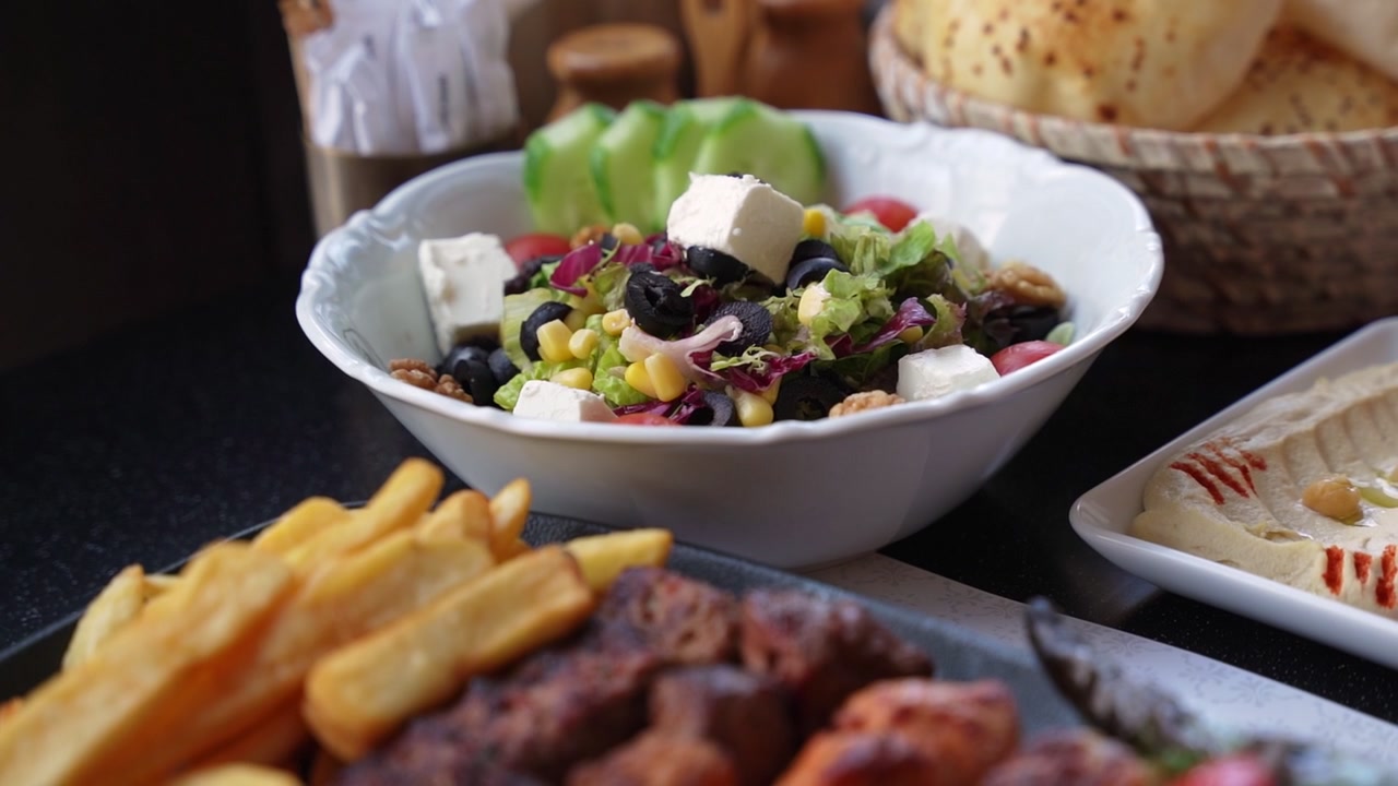 Primo piano di un'insalata fresca e salutare con formaggio feta, olive e mais, servita su un tavolo di ristorante con hummus e spiedini alla griglia in piena luce naturale.