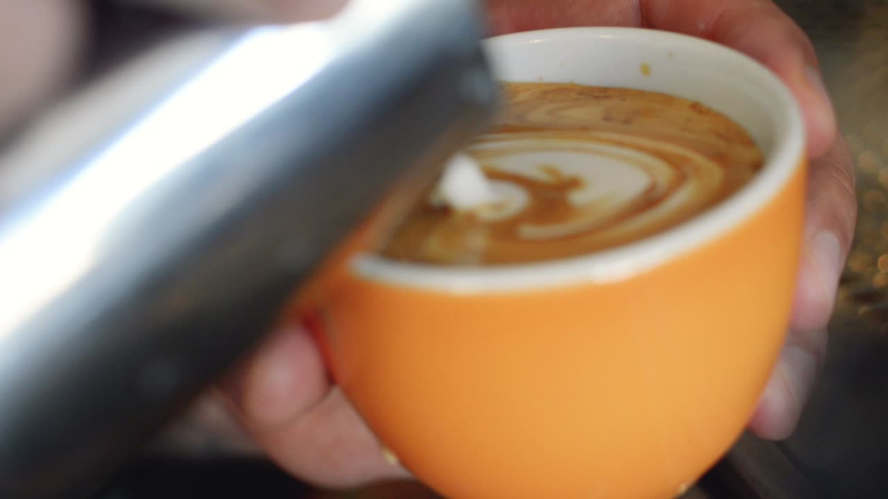 un habilidoso barista sirviendo un latte art con diseño de rosetta, con leche al vapor, en una taza de café naranja | cafetería gourmet | filmado en hd a 24 fps cinemáticos