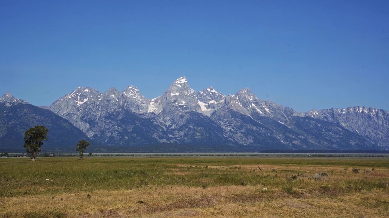 Inquadratura in tilt-up che rivela l'aspra catena montuosa del Teton che si erge sopra un campo di erba secca a Mormon Row nel Parco Nazionale del Grand Teton, catturando un classico paesaggio del Wyoming sotto la luce del sole a luglio